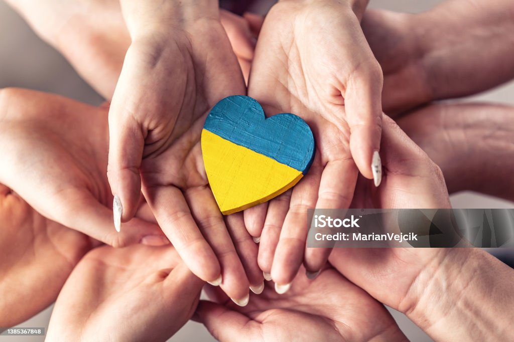 We stand with Ukraine symbol as numerous hands hold one another in unity with a wooden heart painted in Ukrainian flag colors on top. Support Stock Photo