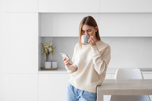 A young beautiful woman using a smartphone and drinking morning tea or coffee in the kitchen at home
