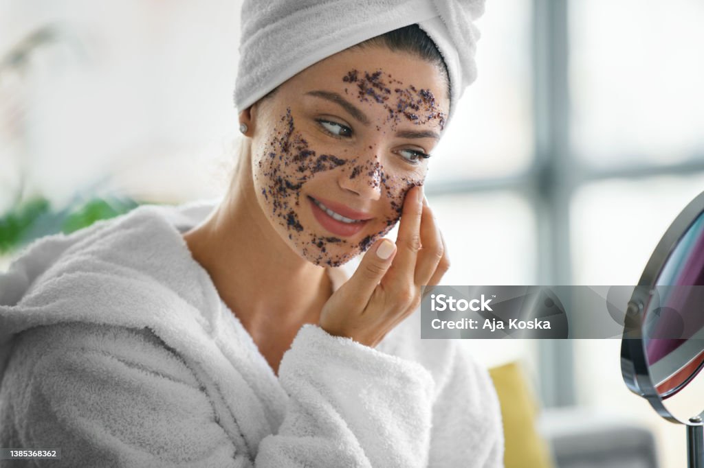 Face scrub for glowing skin. Young woman applying face scrub product on her face while looking at mirror, wearing a bathrobe with her hair wrapped in towel. Exfoliation Stock Photo