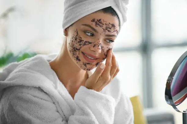 Young woman applying face scrub product on her face while looking at mirror, wearing a bathrobe with her hair wrapped in towel.