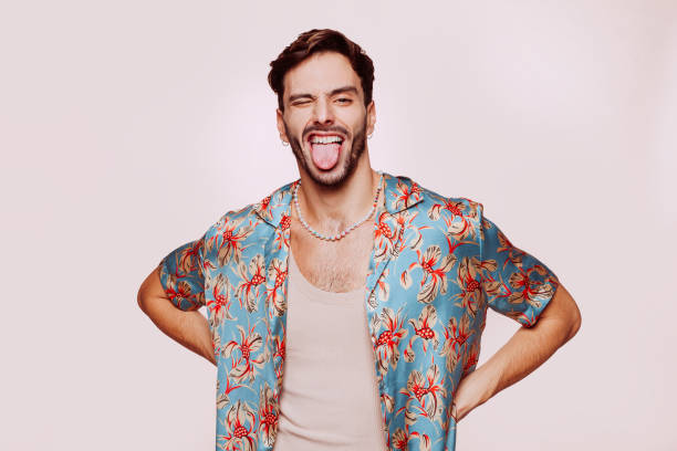 Young handsome homosexual man posing with funny face at studio Portrait of young attractive caucasian homosexual man doing funny face over beige wall. Handsome homosexual in stylish clothes posing with hands on the hips at studio. young man wink stock pictures, royalty-free photos & images