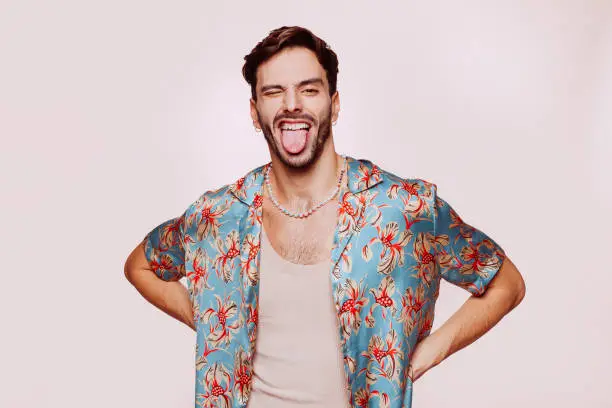 Portrait of young attractive caucasian homosexual man doing funny face over beige wall. Handsome homosexual in stylish clothes posing with hands on the hips at studio.