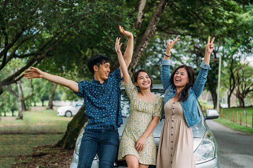 Three Asian friends on a road trip taking a break, leaning on the car waving to camera
