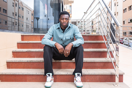 Full body of confident serious young African American male with curly dark hair in trendy outfit sitting on stairs of modern building and looking at camera in city