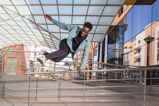 Full body of self assured young positive African American guy with dark hair in trendy outfit jumping over metal railing near modern building on sunny day in city
