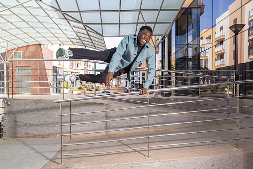Full length of cheerful young black male with dark hair in stylish clothes smiling while jumping over metal railing near contemporary building on city street