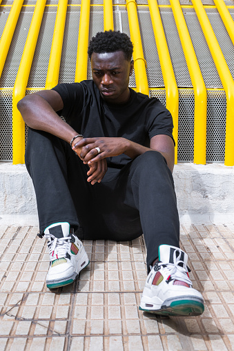 Full body of upset young African American man with curly dark hair in casual clothes and trendy sneakers sitting on ground on street and looking down