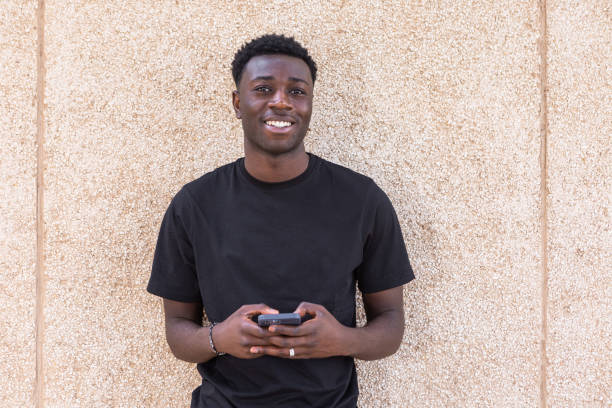 Positive black man using smartphone and smiling at camera on street Cheerful young African American guy with dark curly hair in black t shirt smiling and looking at camera while messaging on smartphone against beige wall on street black man stock pictures, royalty-free photos & images