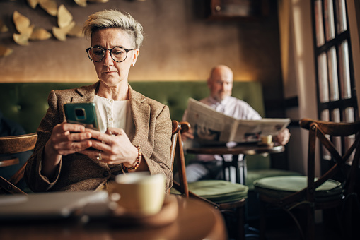 Mature woman using smart phone on coffee break, while man is behind her is reading newspaper in cafe.