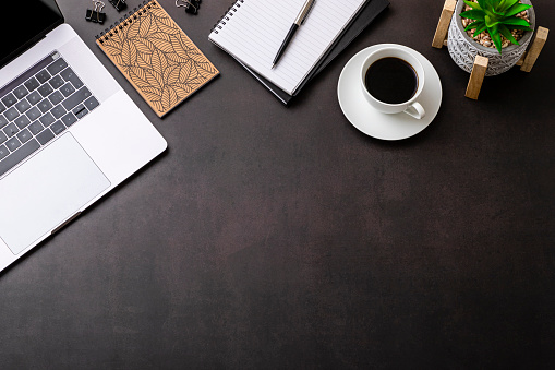 Office desk: Top view of various office supplies such as a laptop, a note pad, a coffee cup and a plant. All the objects are at the top of the image leaving a useful copy space at the bottom on black background.