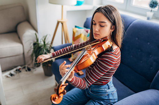 Talented young violinist playing her favorite instrument while sitting on the comfortable sofa of her living room.