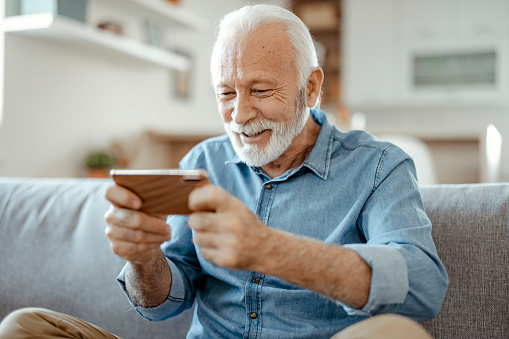 Bearded senior man relaxing on cozy couch with phone, spending leisure time at home, lazy weekend, satisfied male using smartphone