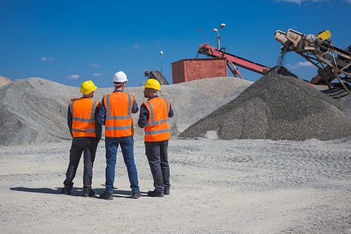 Open-pit mine engineer and workers wearing protective clothes and helmets standing in front of pile of gravel, discussing plans.