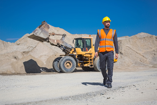 Open-pit mine worker wearing protective clothes and helmet walking in front of pile of gravel and excavator.