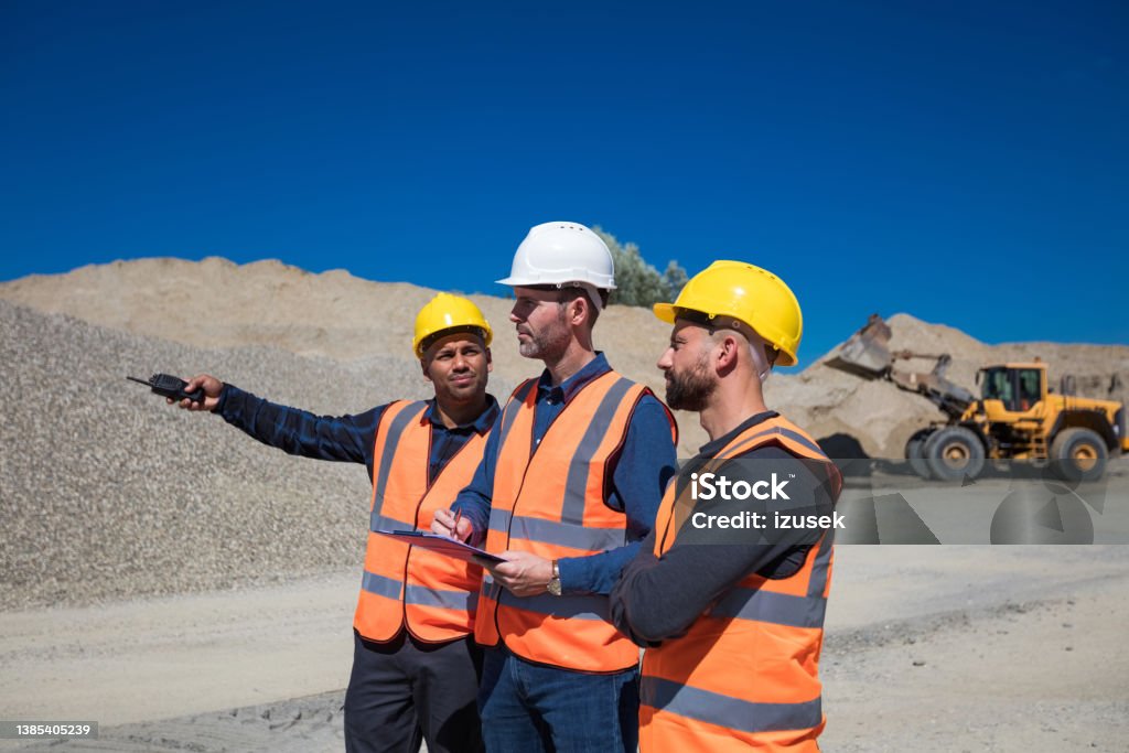 Open-pit mine workers and engineer Open-pit mine workers and engineer wearing protective clothes and helmets discussing in front of pile of gravel. Adult Stock Photo