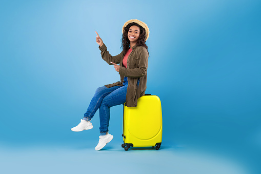 Happy young black woman sitting on bright suitcase and pointing at empty space for travel agency ad on blue studio background. Cheerful female traveler with luggage going on summer vacation