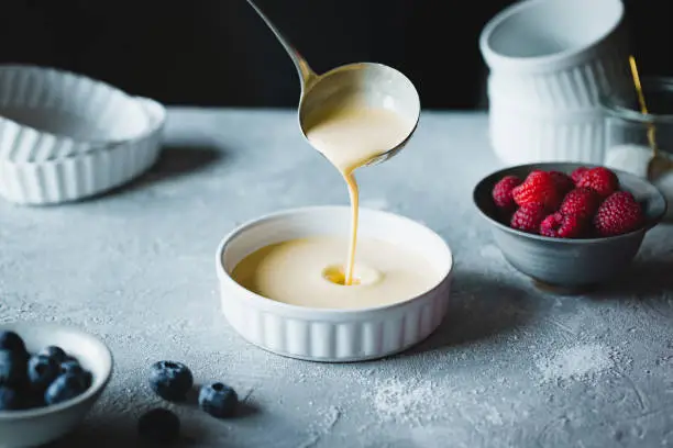 Close-up of chef filling a white plate with custard using ladle and berry fruits in bowls over kitchen table. Chef preparing creme brulee in kitchen.