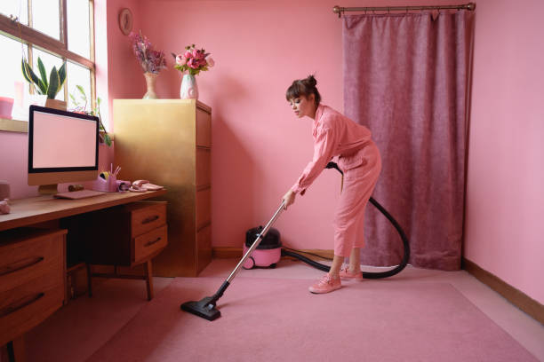 Mature woman vacuuming carpet in pink home office Full length view of multiracial woman with brown hair wearing matching monochrome jumpsuit and doing housecleaning chores. monochrome house stock pictures, royalty-free photos & images