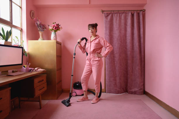 Full length portrait of woman cleaning pink home interior Early 40s multiracial woman in matching monochrome jumpsuit standing in home office with vacuum cleaner, hand on hip, and smiling at camera. offbeat stock pictures, royalty-free photos & images
