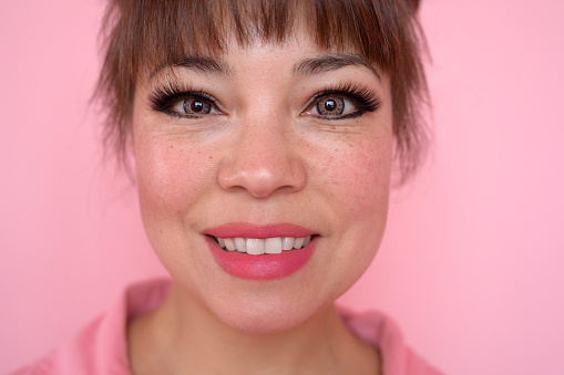 Freckle-faced woman with brown hair up wearing pink open collar garment, matching lipstick, and smiling at camera against pastel background.