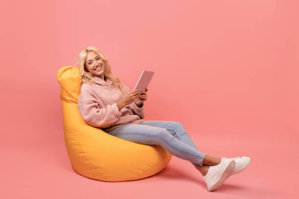 Happy lady using digital tablet while sitting in beanbag chair over pink background with free space