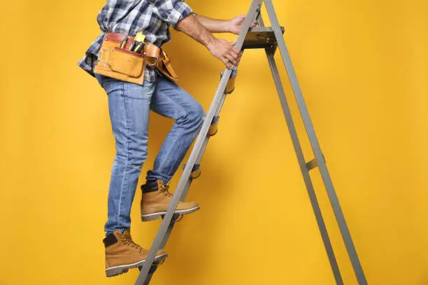 Photo of Professional builder climbing up metal ladder on yellow background, closeup