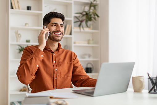 Handsome young Arabic independent contractor working online from home office, sitting in at desk using laptop and having phone conversation with business partner, discussing plan, free copy space