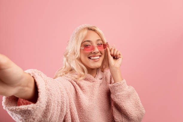 mulher alegre e elegante fazendo selfie, usando óculos coloridos e capuz, sorrindo para a câmera sobre fundo rosa - pink glasses - fotografias e filmes do acervo