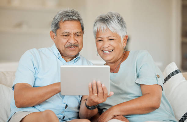 shot of a happy senior couple relaxing and using a tablet on the sofa at home - senior adult happiness computer looking at camera imagens e fotografias de stock