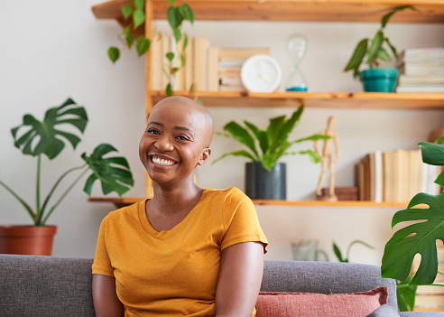 A young woman smiles while sitting on the couch in front of a bookshelf filled with plants. High quality photo