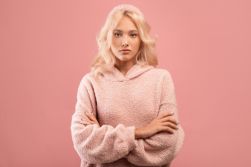 Serious stylish lady looking at camera and posing with folded arms over pink studio background. Confident woman crossing hands, thinking about something