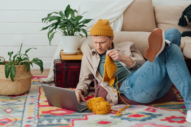 Young serious man looking at laptop. Man learning new hobby, knitting on needles. Knitting project in progress. stock photo