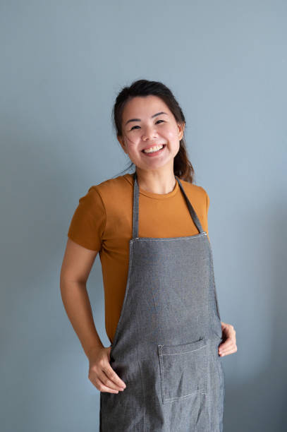 Mid adult Asian woman barista small business owner in apron looking at camera and standing against gray background.