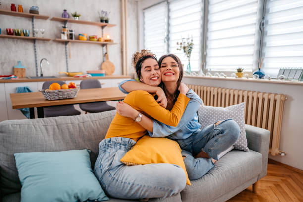 Two Female Friends Embracing Each Other Two beautiful female friends sitting on the sofa in the living room and embracing each other. female-friendship stock pictures, royalty-free photos & images