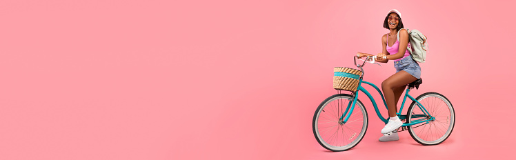 Full length of lovely black woman in casual summer clothes riding bicycle over pink studio background. Positive African American lady on bike going cycling, panorama with copy space, banner