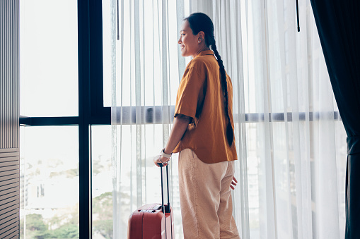 A smiling Asian tourist looking out the hotel room window and holding her suitcase.