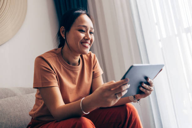 femme thaïlandaise heureuse assise sur le canapé et lisant un roman passionnant sur sa tablette - thai ethnicity photos et images de collection