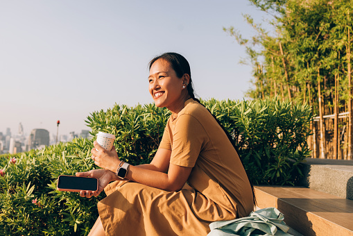 A smiling Asian woman sitting on the stairs in the park and drinking a cup of coffee on a sunny day.