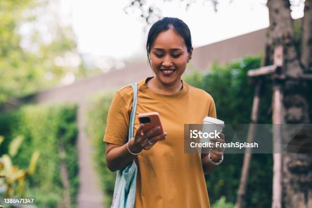 Portrait Of A Smiling Asian Woman In A Casual Yellow Dress Checking Her Mobile Phone As She Walks In The Park Carrying A Cup Of Coffee Stock Photo - Download Image Now