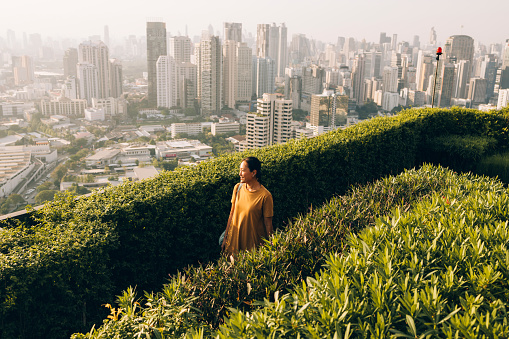 A happy Thai woman walking in the park with the cityscape behind her.