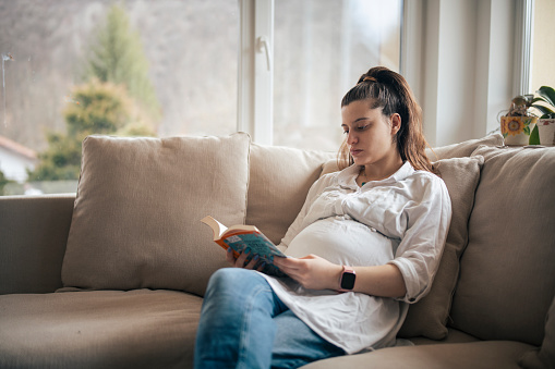 Young pregnant woman at home sitting on the sofa and reading a book.