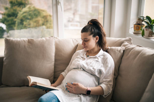 istock Pregnant woman reading a book while relaxing on couch at home. 1385480735