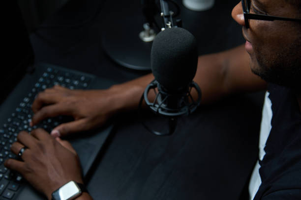 Man with headphones records or broadcasts a podcast using a microphone in studio Close-up of an African podcaster blogger smiling during broadcast of his live audio podcast in studio. Male radio host with glasses, using a microphone and headphones, conducts a podcast or interview radio dj stock pictures, royalty-free photos & images