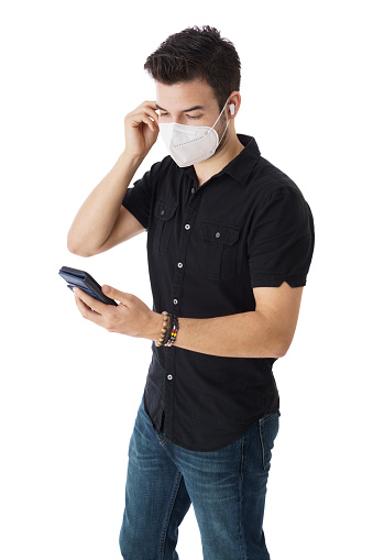 A young latin man looking at his mobile phone and fixing his face mask.