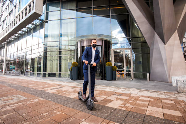 Casual businessman with a protective face mask seen standing with his electric scooter in front of a hotel after finishing work day during coronavirus pandemic.
