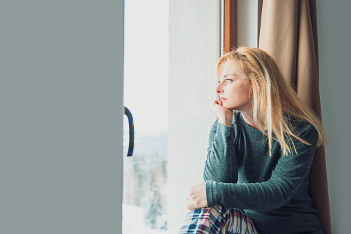 Pensive Blonde Woman sitting on windowsill looking out window. Copy space