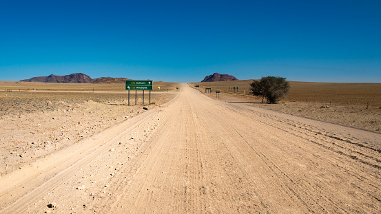 road trip in Namibia - endless straight gravel road washboard in the plains of Namib Naukluft Park - intersection of two gravel roads - green road sign Solitaire-Windhoek