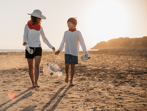 a mother and her son, walking along the beach at sunset, while collecting plastic bottles from the shore. concept care of the planet and ecology.