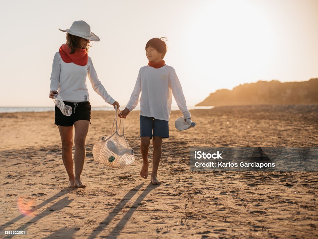 a mother and her son, walking along the beach at sunset, while collecting plastic bottles from the shore. concept care of the planet and ecology. Eco Tourism Stock Photo