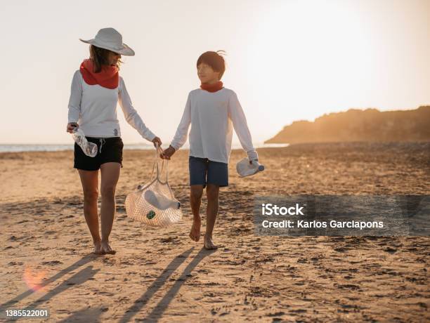 A Mother And Her Son Walking Along The Beach At Sunset While Collecting Plastic Bottles From The Shore Concept Care Of The Planet And Ecology Stock Photo - Download Image Now